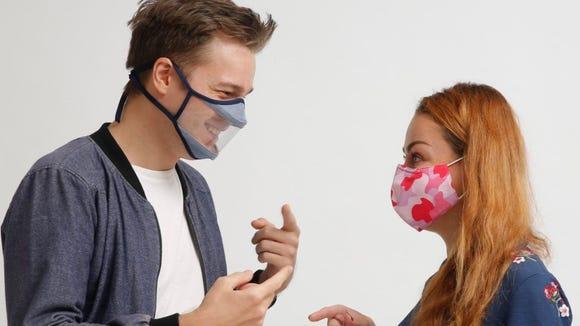 A young man wearing a clear transparent mask is smiling and talking to a young woman in a regular mask who is looking at him